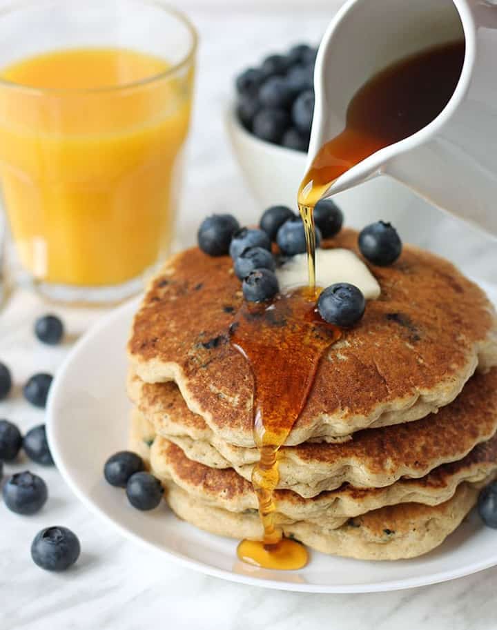 a stack of vegan blueberry pancakes on a white plate with a glass of orange juice ein the background and maple syrup being poured over the pancakes 