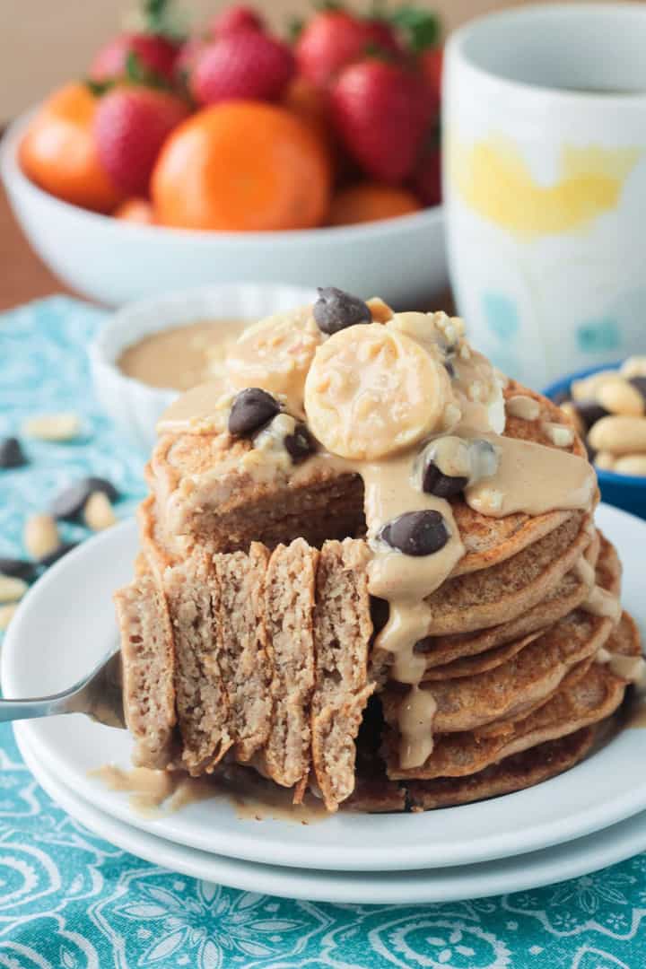 a stack of vegan peanut butter pancakes on a white plate with a fork and a bowl with fruit in the background
