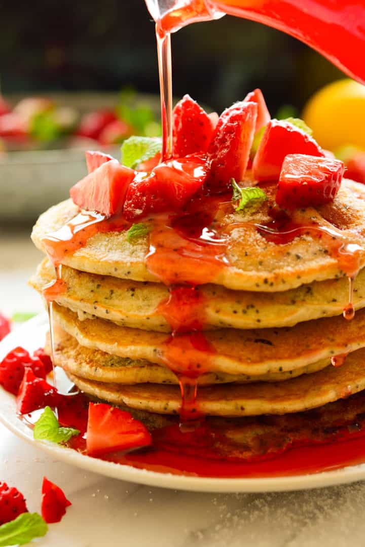 a stack of vegan pancakes on a red plate with strawberries on top and strawberry sauce being poured over the pancakes