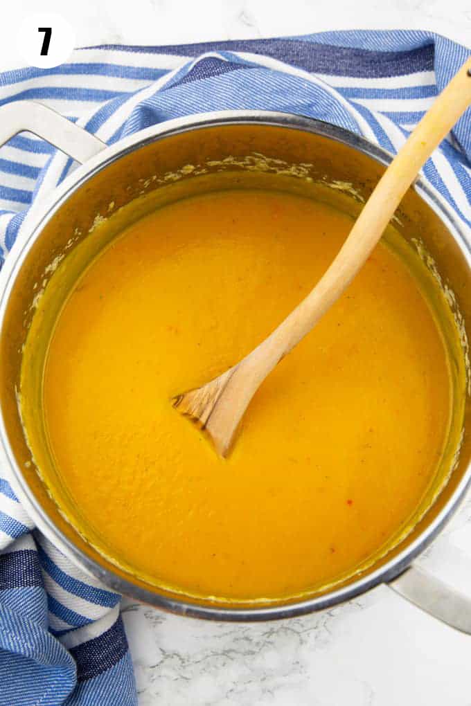 a pot with vegan pumpkin soup on a marble countertop with a wooden spoon 