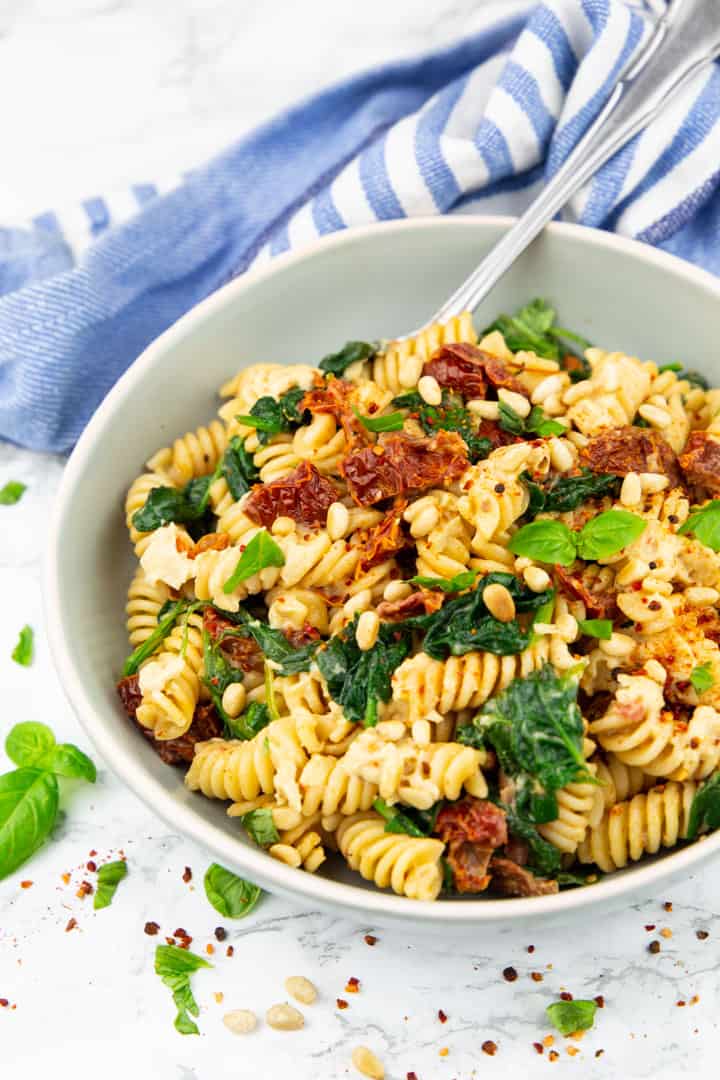 a bowl with rotini pasta with spinach, sun-dried tomatoes, and hummus on a marble countertop 