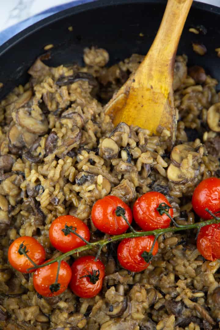 a close-up photo of mushroom risotto with roasted tomatoes in a black pan with a wooden spoon