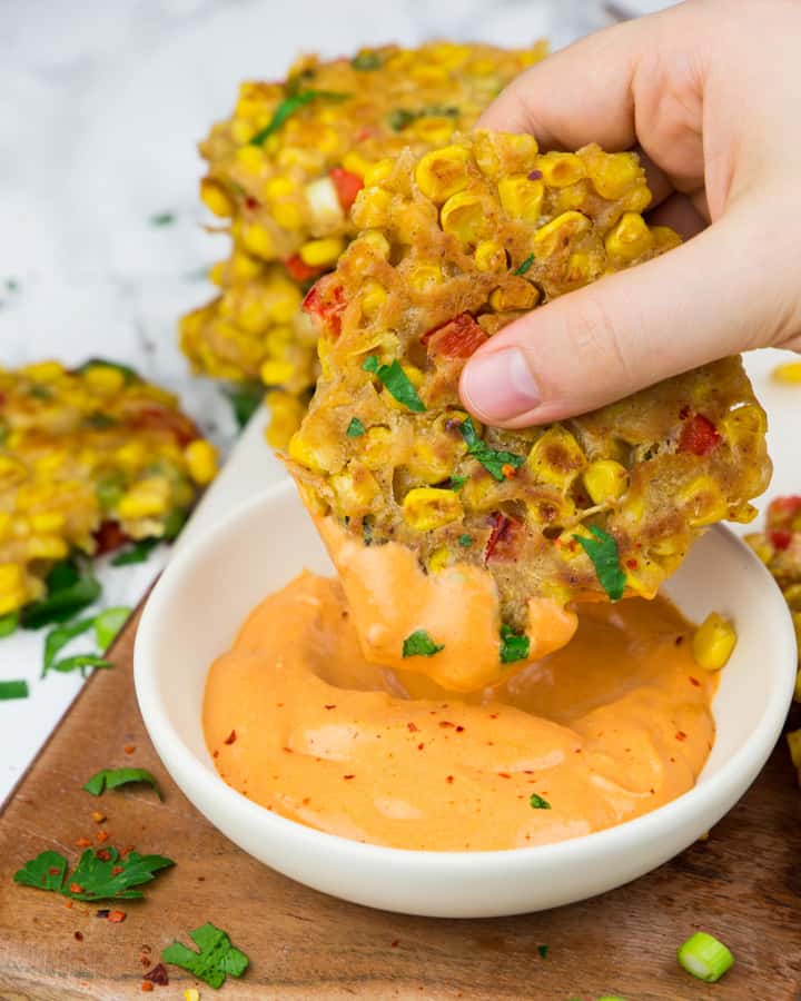 a hand dipping a corn fritter into a small bowl of paprika mayonnaise with more fritters in the background