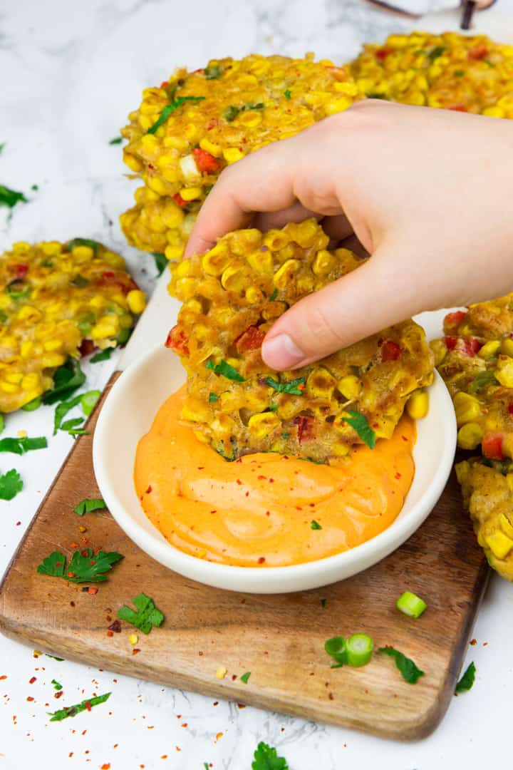 a hand dipping a corn fritter into a bowl of paprika dipping sauce on a wooden board with chopped green onions and parsley on the side 