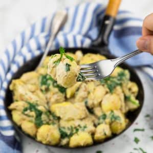 Spinach Artichoke Gnocchi in a black pan on a marble countertop with a hand picking up gnocchi with a fork