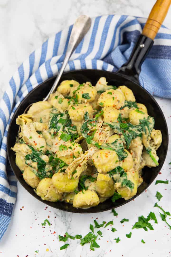 spinach artichoke gnocchi in a black pan with a spoon on a marble countertop with a blue and white tablecloth in the background