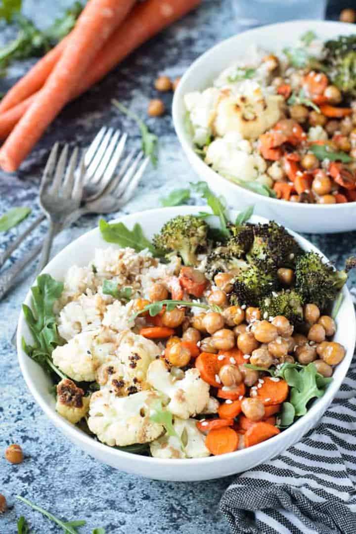 two bowls with roasted cauliflower, broccoli, carrots and chickpeas on a blue countertop 