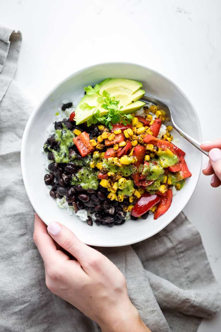 a hand holding a white bowl with rice, black beans, bell pepper, and avocado over a white countertop 