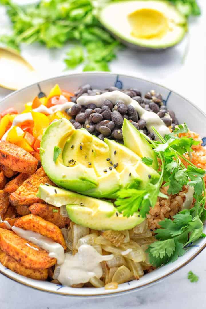 a white bowl with sweet potatoes, black beans, and avocado on a white countertop with an avocado and fresh cilantro in the background