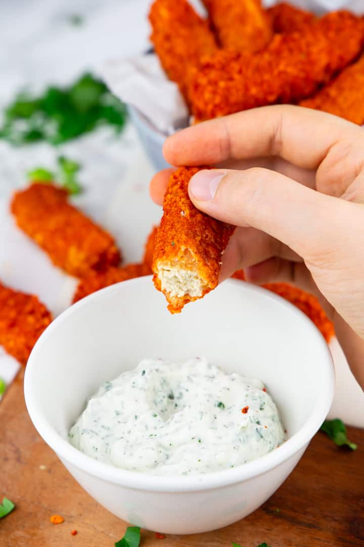 a hand holding a tofu stick over a bowl with vegan ranch with more tofu fingers in the background 