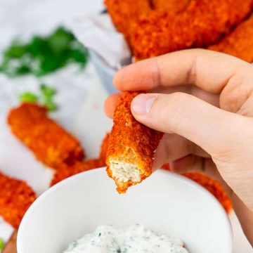 a hand holding a tofu stick over a bowl with vegan ranch with more tofu fingers in the background