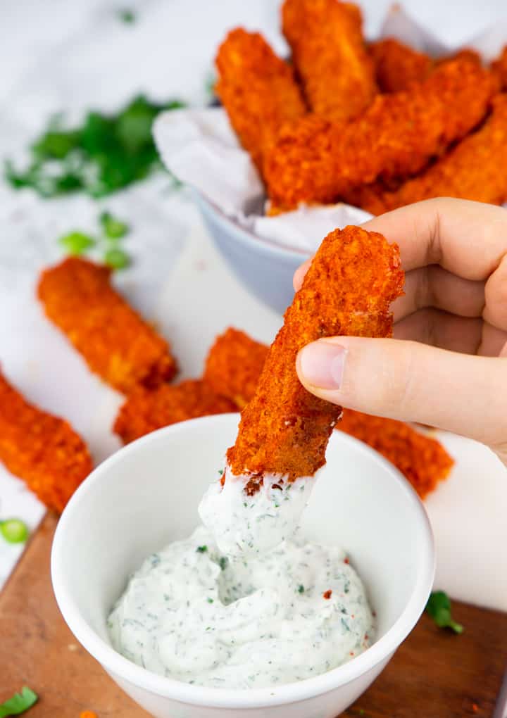 a hand dipping a tofu stick into a bowl of vegan ranch sauce with more tofu sticks in the background 