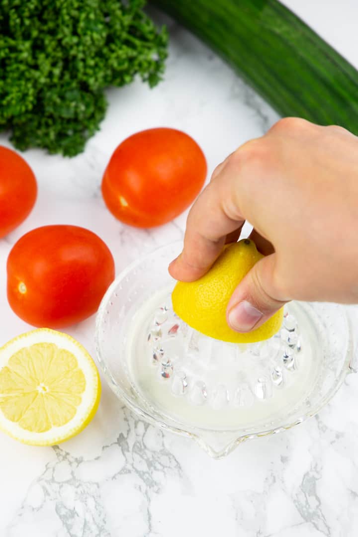 a hand juicing a lemon with three tomatoes, a cucumber, and parsley in the background 