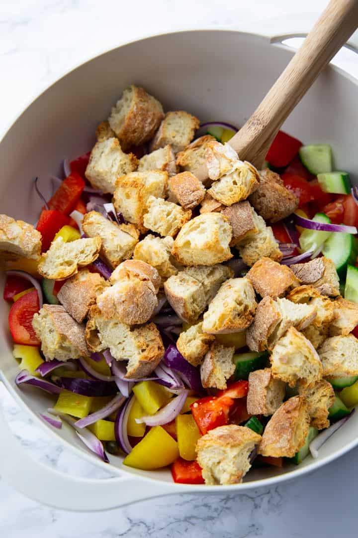 a large grey bowl with chopped tomatoes, cucumber, bell pepper, red onion, and roasted ciabatta cubes on a marble countertop 