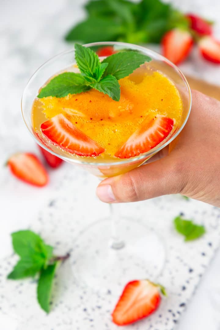a hand holding a glass of champagne and strawberry sorbet with strawberry halves and mint