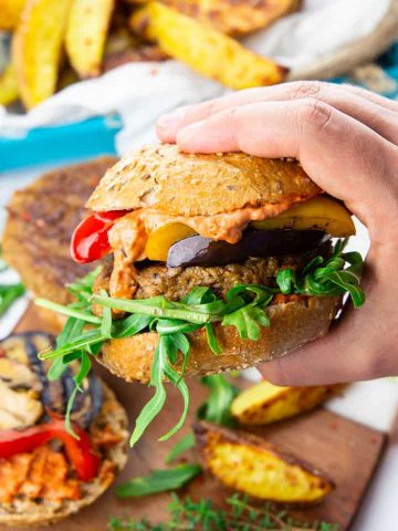 a hand holding a vegan burger with grilled vegetables and arugula with fries in the background