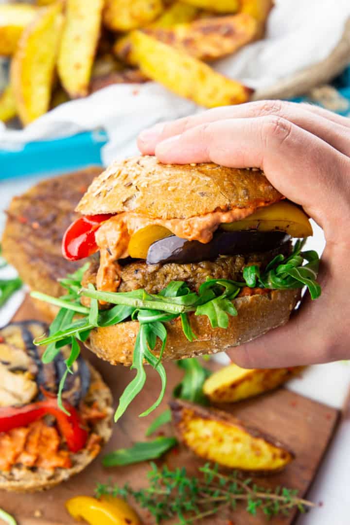 a hand holding a vegan burger with grilled vegetables and arugula with fries in the background 