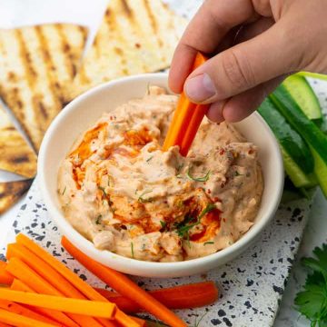 a hand dipping a carrot stick into a small bowl of jackfruit buffalo dip with cucumber and carrot sticks on the side