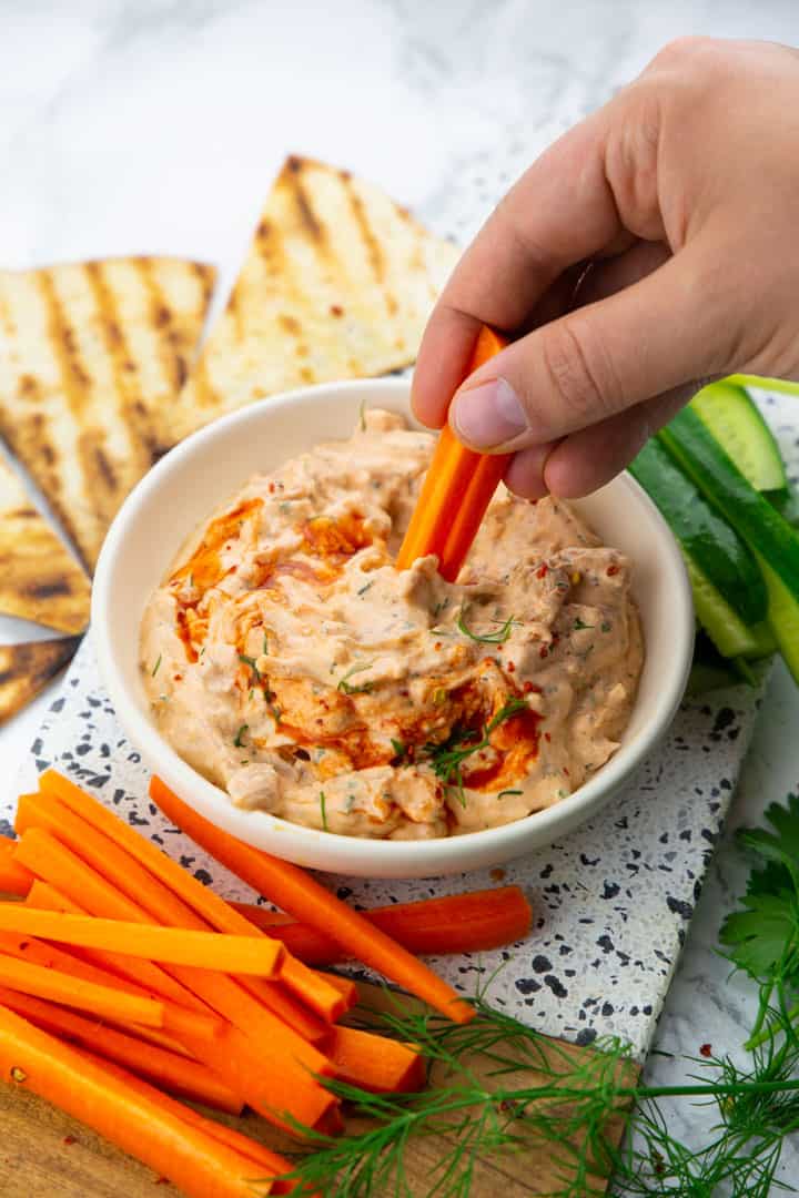 a hand dipping a carrot stick into a small bowl of jackfruit buffalo dip with cucumber and carrot sticks on the side 