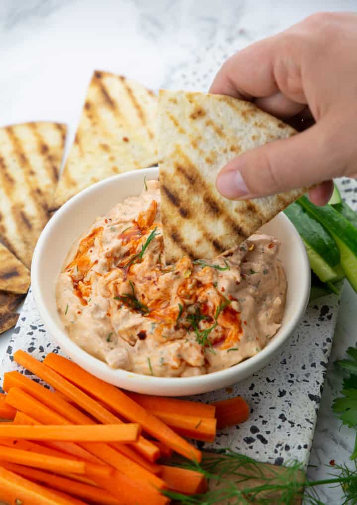 a hand dipping a grilled tortilla triangle into a small bowl with Buffalo jackfruit dip with carrot and cucumber sticks on the side 