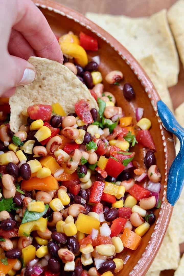 bean salad in a brown bowl with a hand dipping a nacho into the salad 