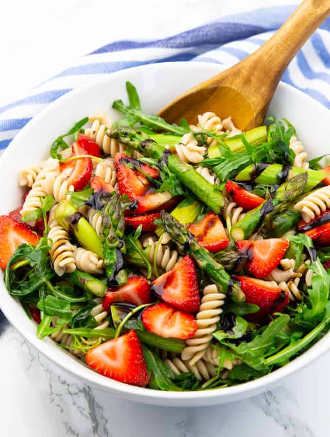 pasta salad with asparagus, strawberries, and arugula in a white bowl with a wooden spoon on a marble countertop 