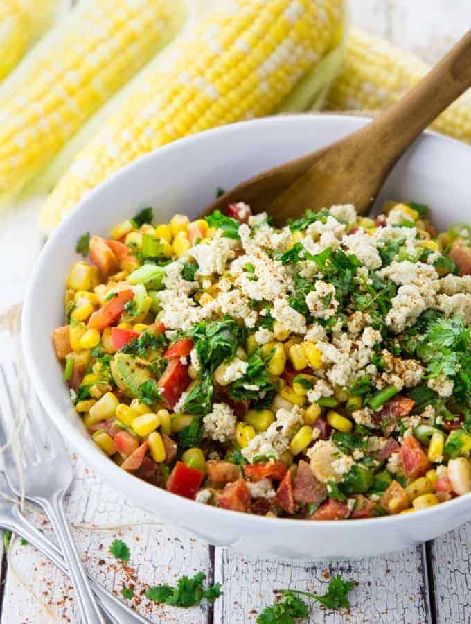 corn salad in a white bowl with a wooden spoon whit four corncobs in the background