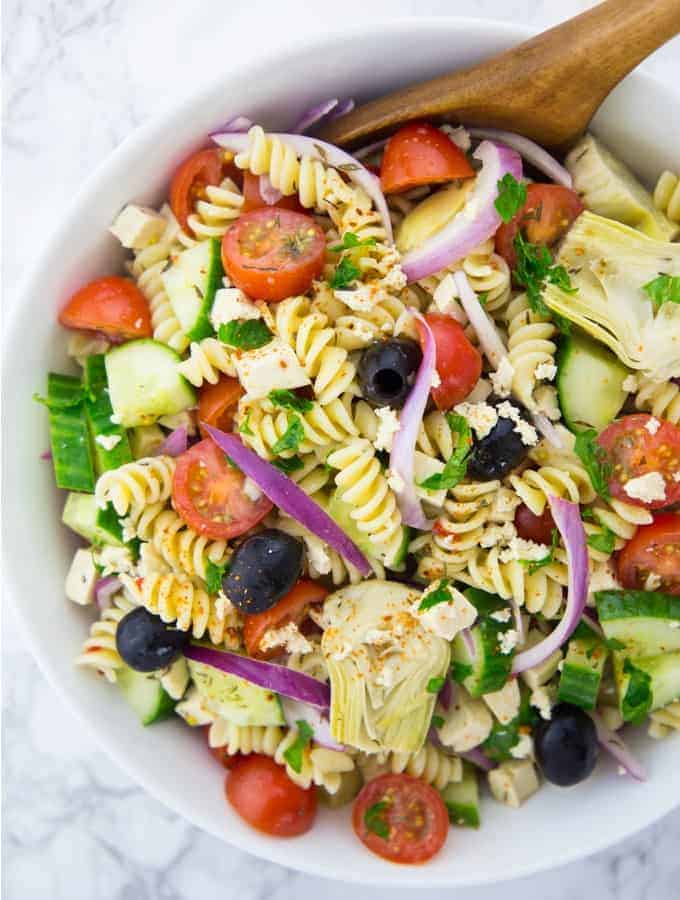 Greek pasta salad with cherry tomatoes, olives, and artichokes in a white bowl with a wooden spoon on a marble countertop 