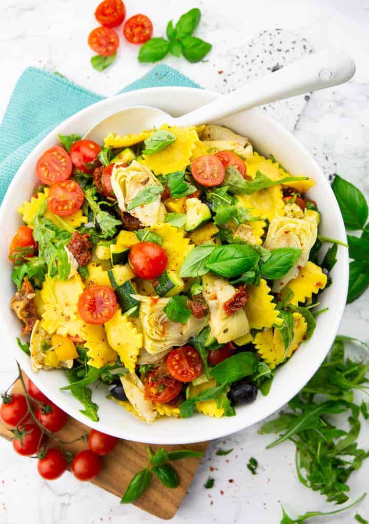 Raviolo salad with artichokes, tomatoes, and zucchini in a white bowl with a white spoon on a marble countertop with tomatoes, basil, and arugula on the side 