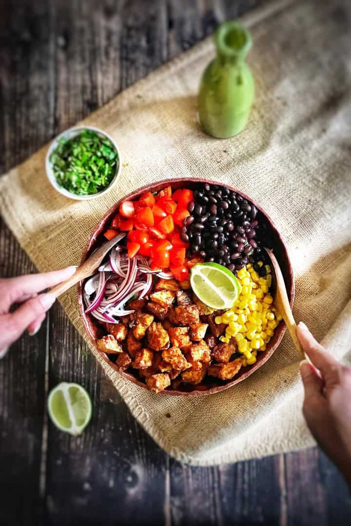 a bowl with Southwestern Salad with Chile Lime Tofu Nuggets on a wooden countertop with a lime and a bottle of dressing on the side