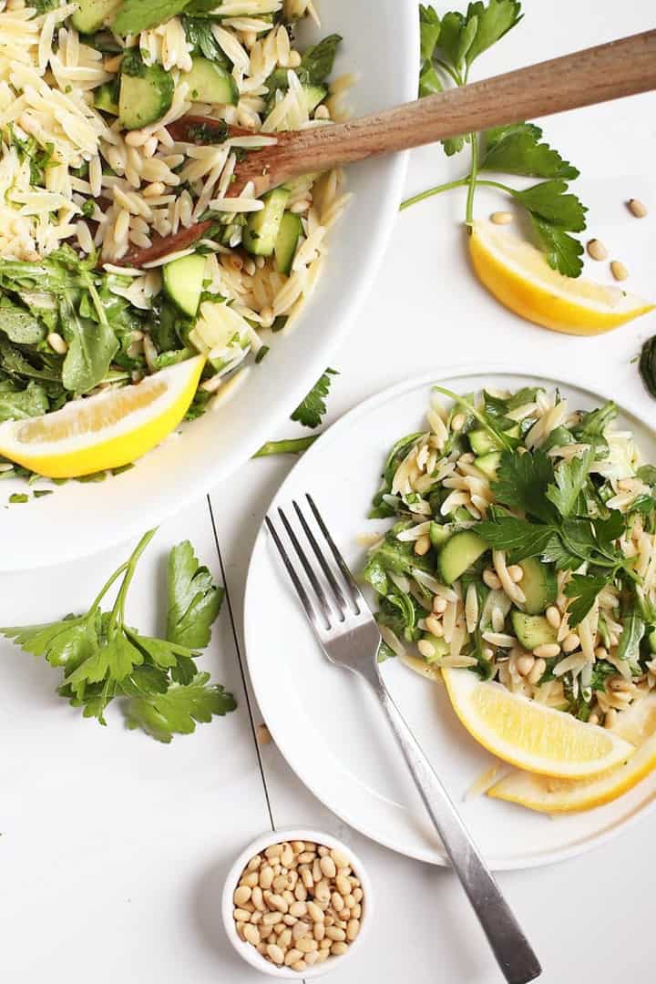 a white plate with lemon orzo salad with a fork and a large bowl with salad in the background 