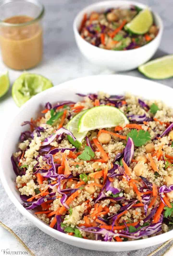 Thai quinoa salad with carrots and red cabbage in a white bowl with another smaller white bowl and limes as well as a glass of peanut dressing in the background