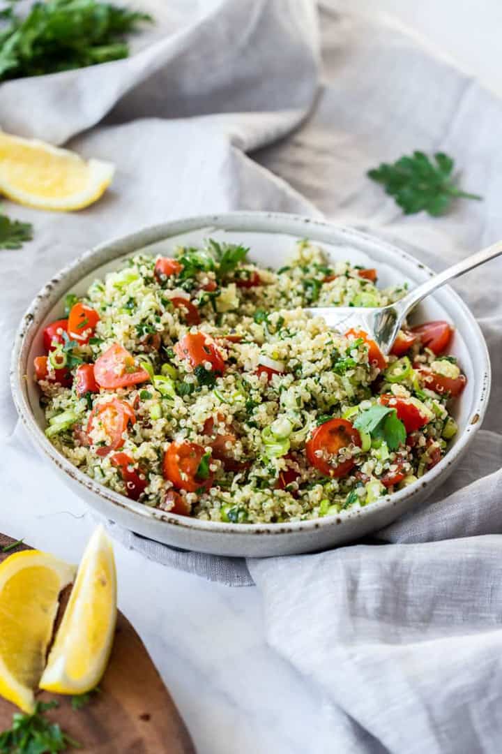 a white bowl with Quinoa Tabouli on a white table cloth with lemon wedges and herbs on the side