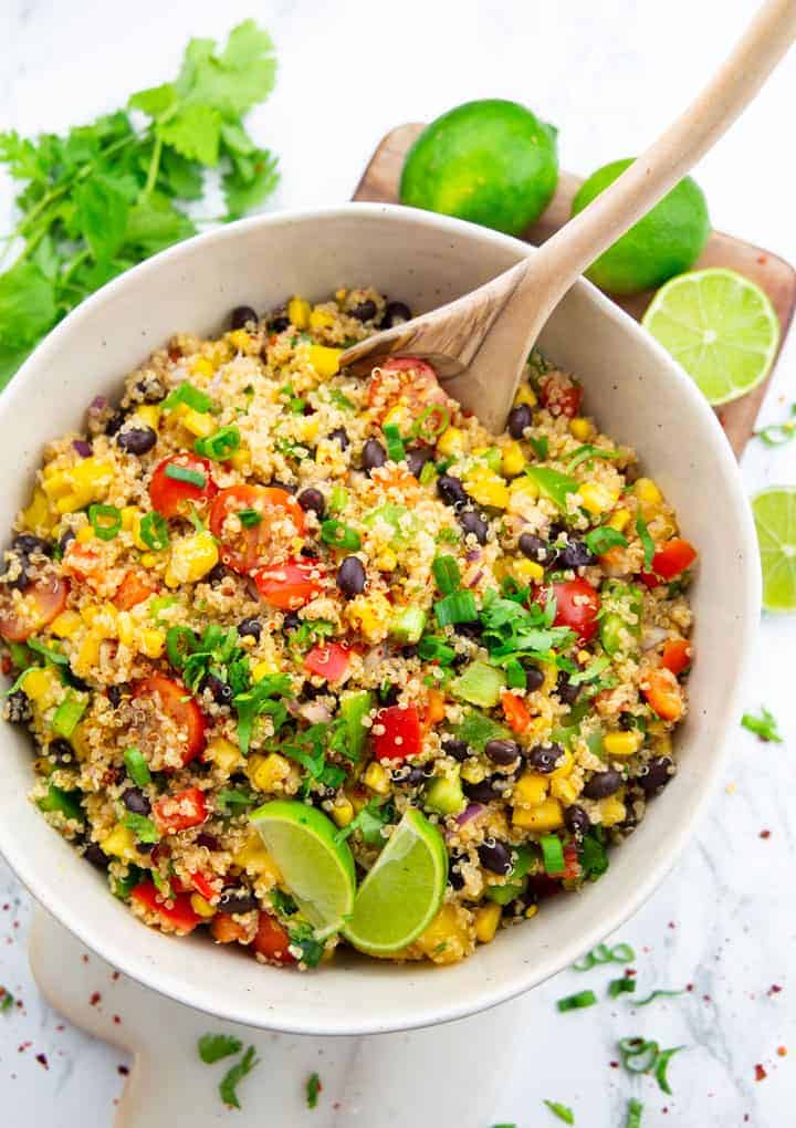 Quinoa Black Bean Salad in a white bowl with a wooden spoon on a marble countertop with three limes in the background 