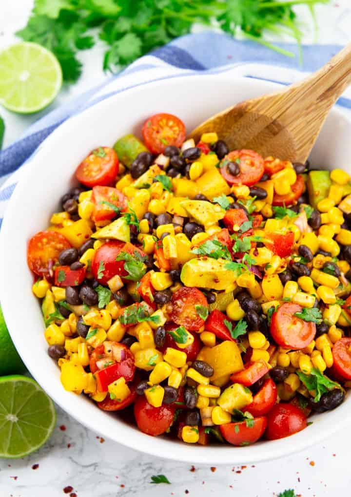 Mexican Salad with corn, black beans, and tomatoes in a white bowl with a wooden spoon on a marble countertop with limes and cilantro in the background 