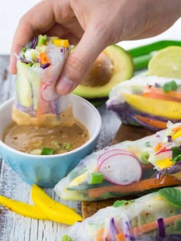 a hand dipping a rice paper roll into a bowl of peanut sauce
