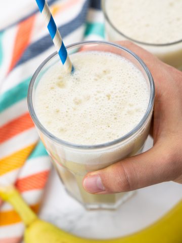 a hand holding a glass with banana milk and a blue straw