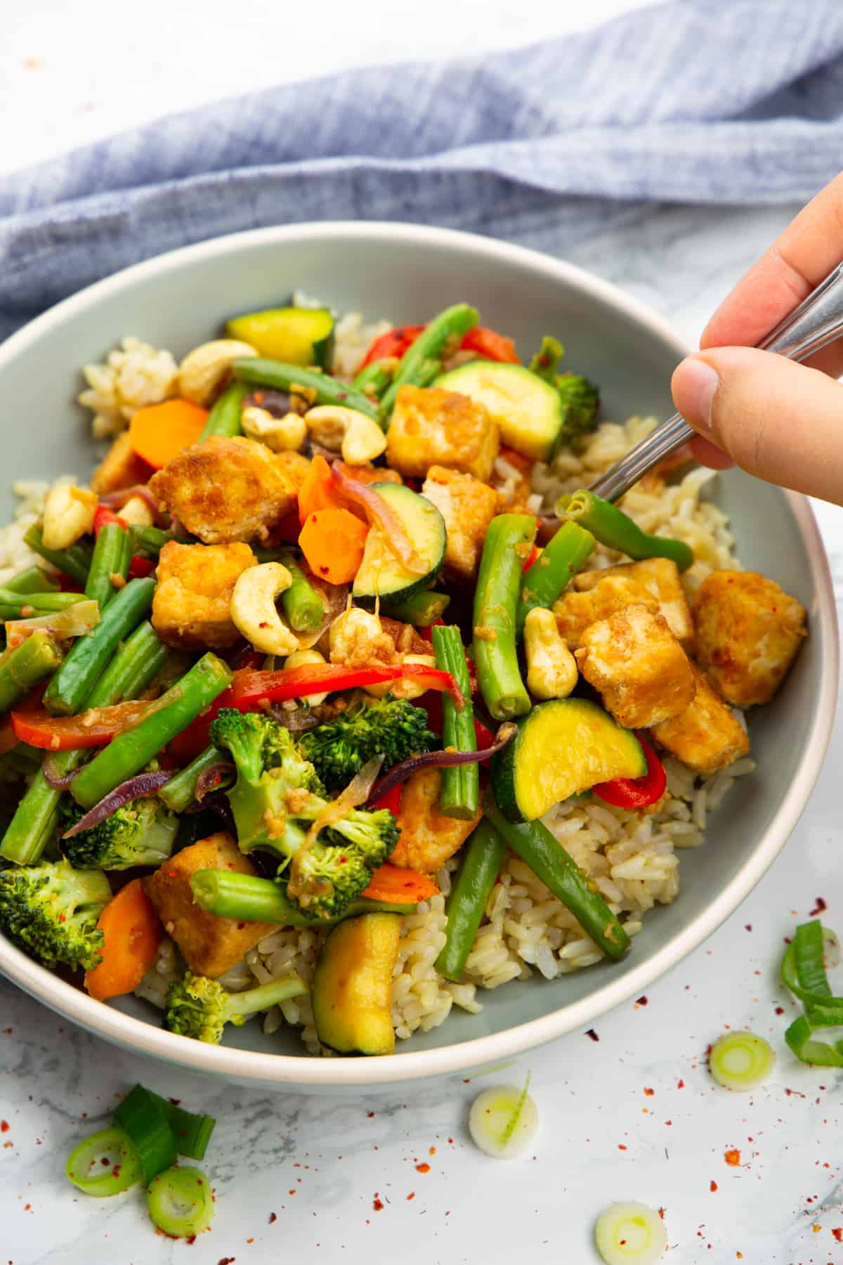 a grey bowl with brown rice and tofu stir fry with a hand holding a fork