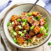 general tso tofu over rice sprinkled with sesame seeds and green onions in a grey bowl on a wooden board