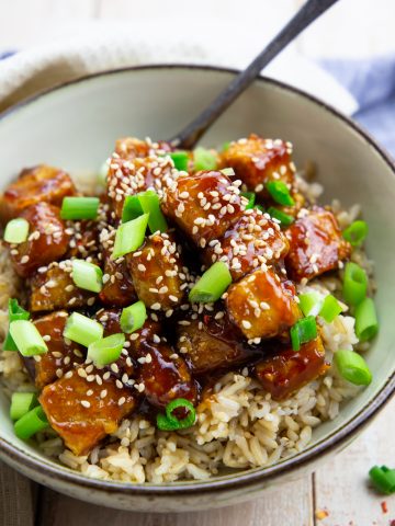 general tso tofu over rice sprinkled with sesame seeds and green onions in a grey bowl