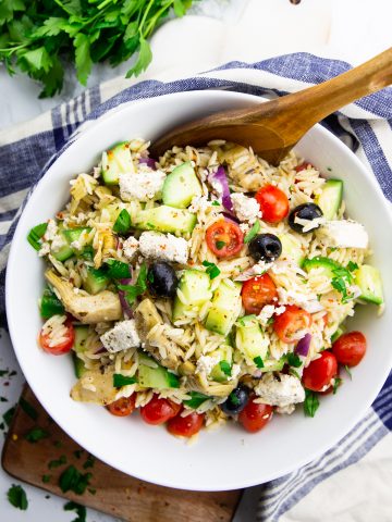 orzo pasta salad in a white bowl with a wooden spoon on a marble countertop