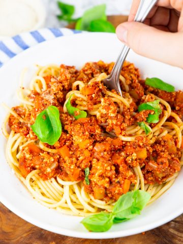 a hand with a fork picking up spaghetti with vegan bolognese out of a white bowl