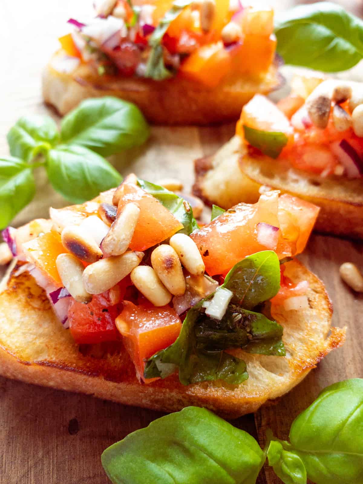 three bruschettas topped with roasted pine nuts on a wooden board with basil on the side