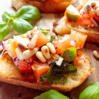 three bruschettas topped with roasted pine nuts on a wooden board with basil on the side