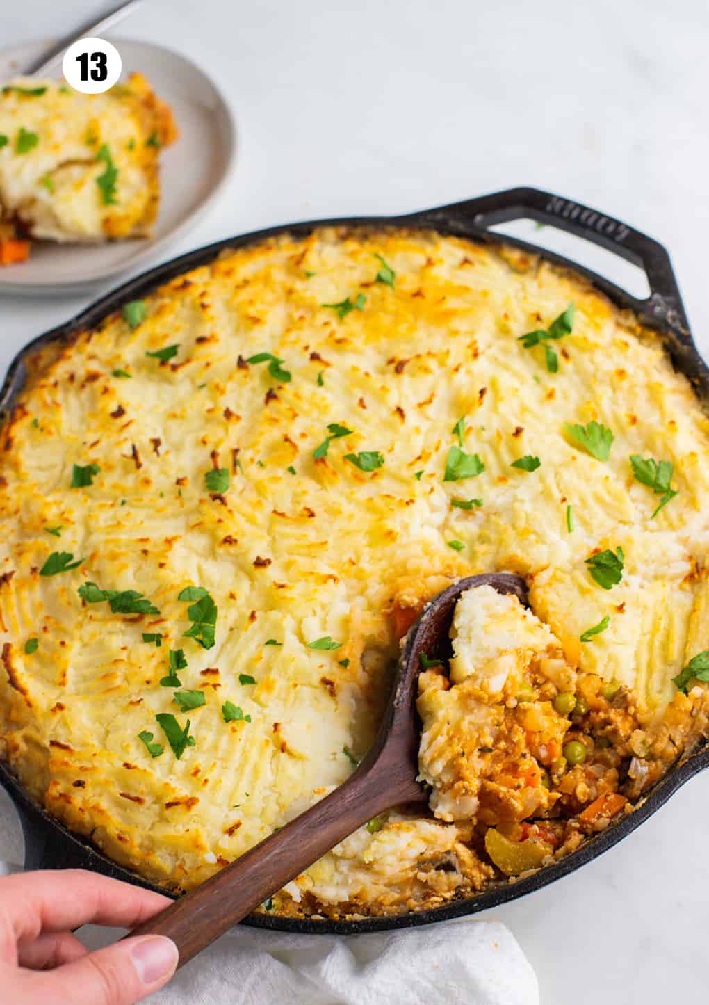 a vegan shepherd's pie in a cast iron pan with a hand spooning some out with a large wooden spoon