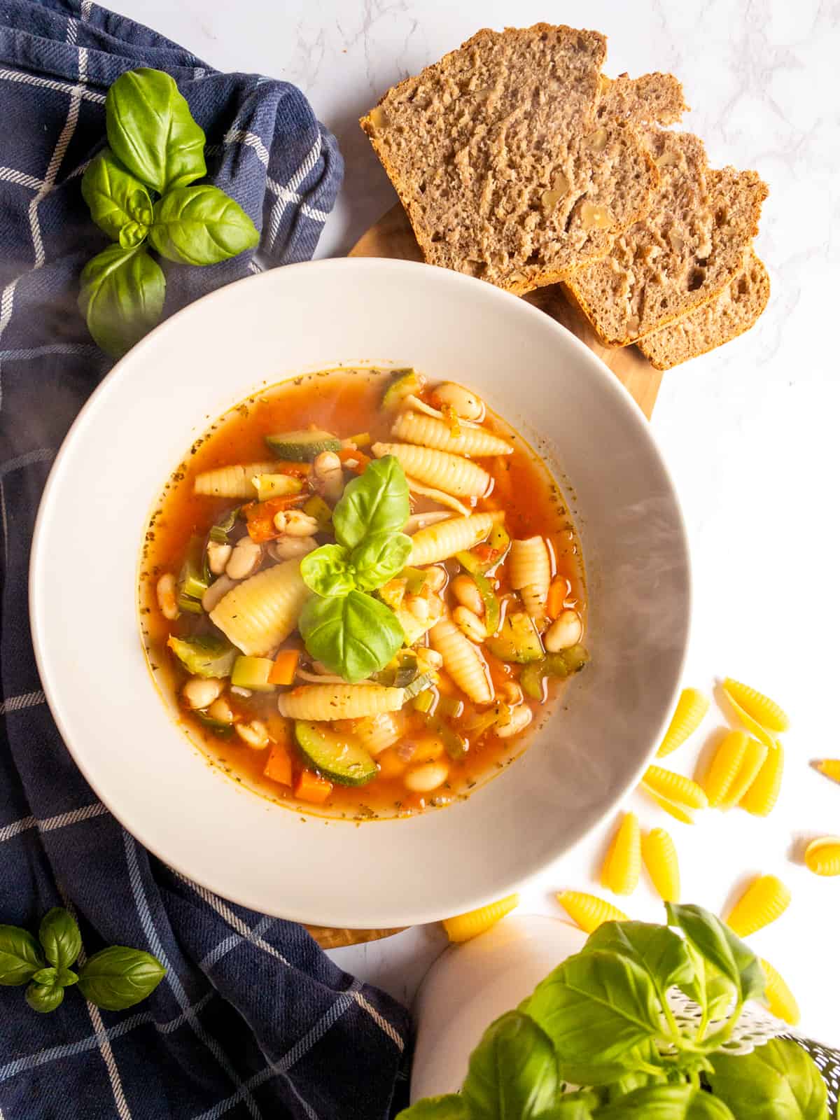 vegan minestrone soup with pasta shells in a white bowl on a white countertop with bread on the side