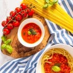 a small bowl with cherry tomato sauce on a wooden board with spaghetti and basil on the side