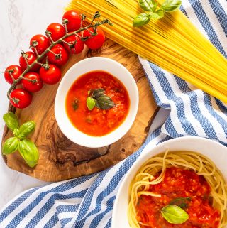 a small bowl with cherry tomato sauce on a wooden board with spaghetti and basil on the side
