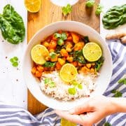 a hand holding a white bowl of sweet potato curry with rice over a wooden board