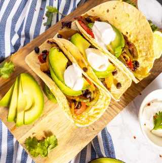 three vegetarian fajitas with sour ceeam and avocado slices on a wooden chopping board
